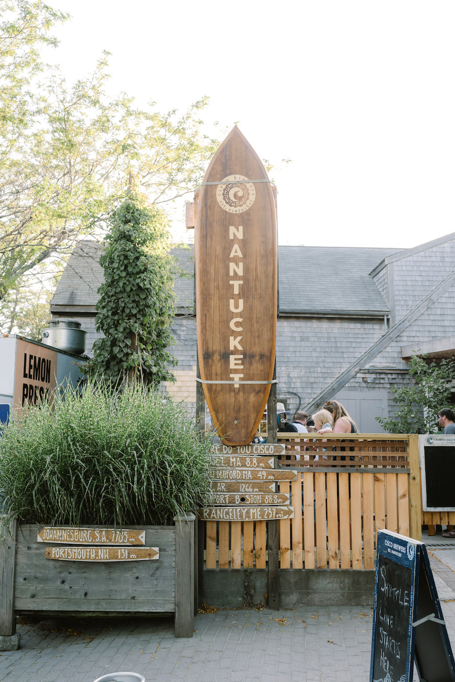 A large wooden surfboard sign reading "Nantucket" stands upright in front of a building with greenery and a wooden fence nearby.
