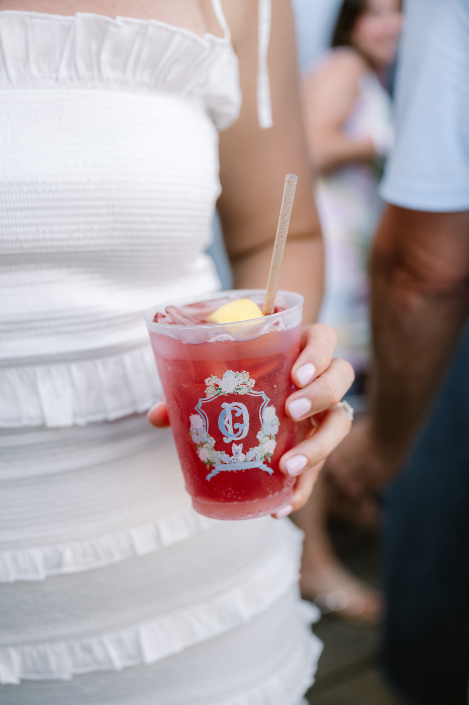 A person in a white ruffled dress holds a red drink with a lemon garnish in a cup featuring a decorative emblem.