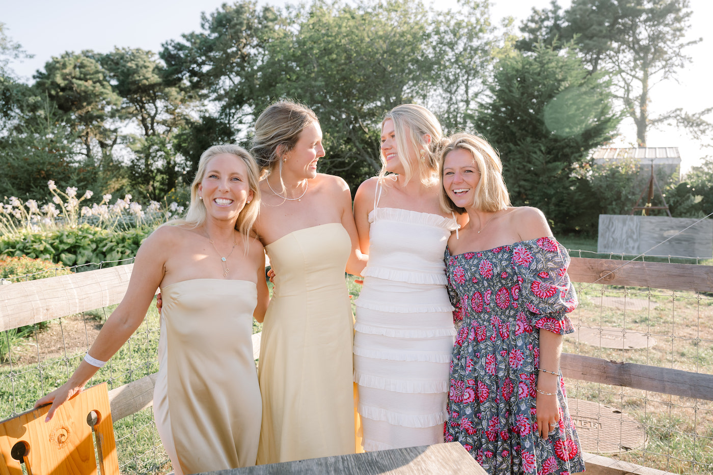 Four women stand outdoors near a wooden fence, dressed in light-colored summer dresses, smiling and posing together in natural sunlight.
