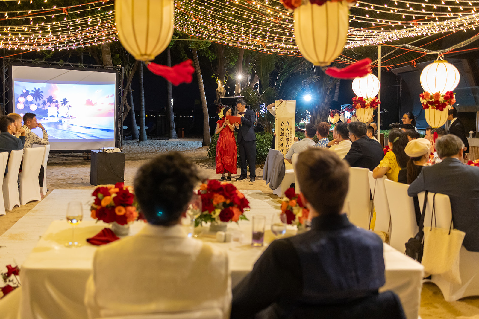 Guests seated at decorated tables watch a speaker on stage during an outdoor evening event with string lights and large lanterns overhead.