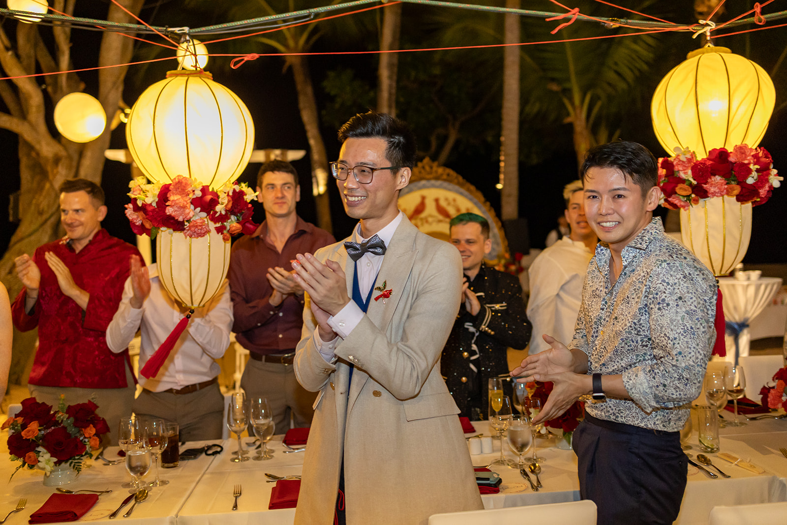 A group of people stand and clap around a decorated dinner table outdoors at night, with large lanterns and floral arrangements in the background.