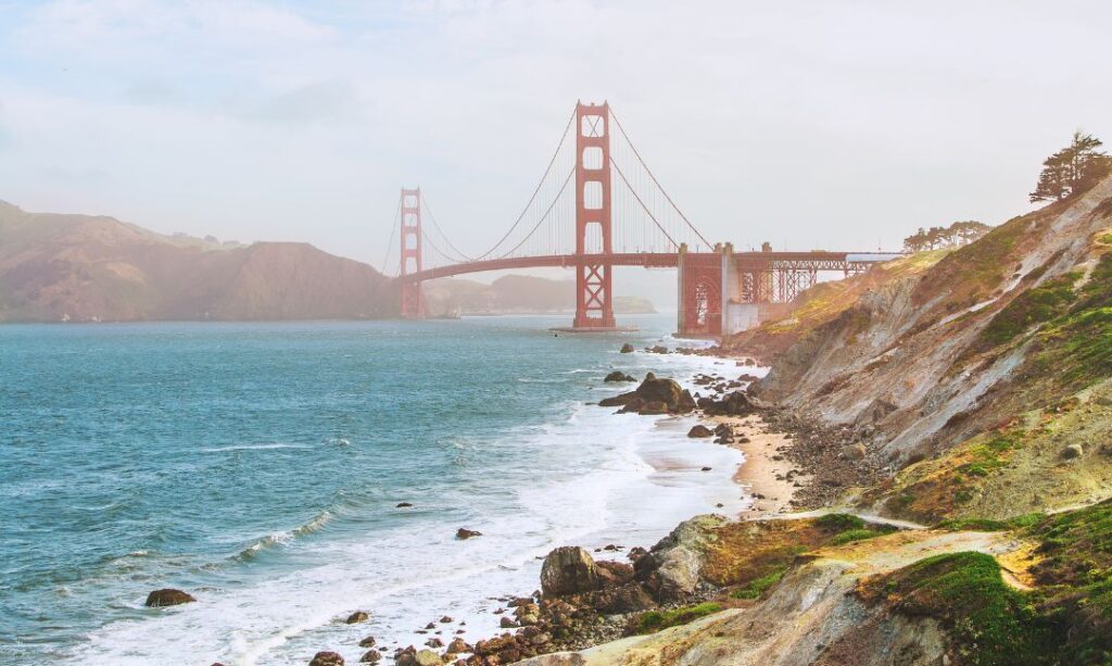 Golden Gate Bridge spans across the water with waves crashing along a rocky shoreline under a cloudy sky.