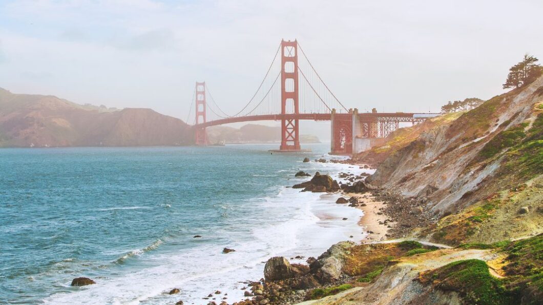 Golden Gate Bridge spans across the water with waves crashing along a rocky shoreline under a cloudy sky.