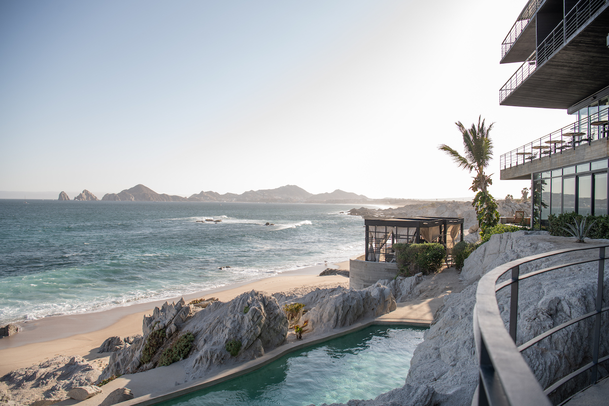 View of a beachfront property with a pool, rocky shoreline, palm tree, and ocean waves; distant mountains are visible under a clear sky.