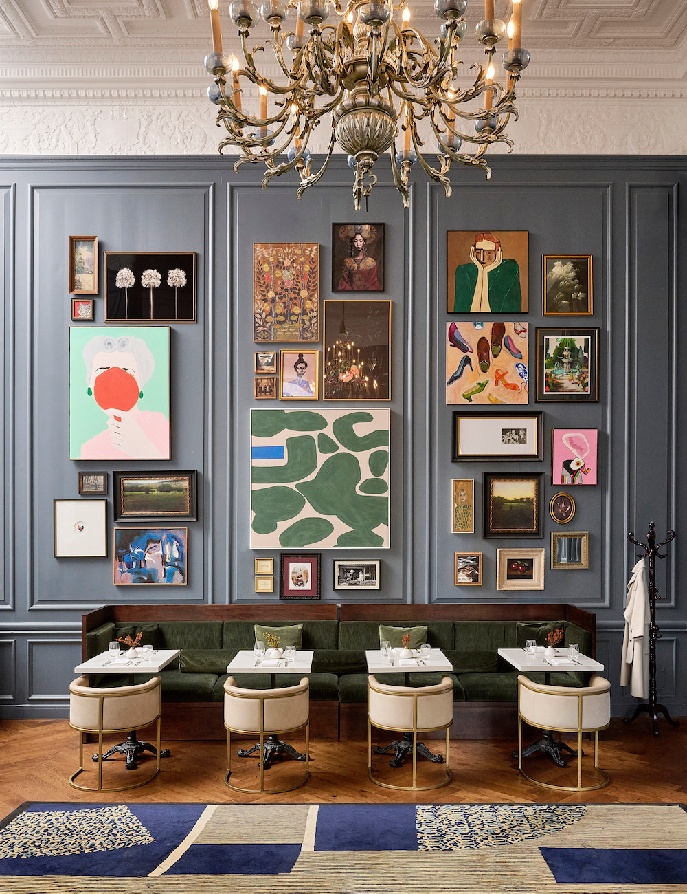A stylish dining area with a green velvet bench, four white chairs, set tables, a large chandelier, and a gallery wall of assorted framed art.