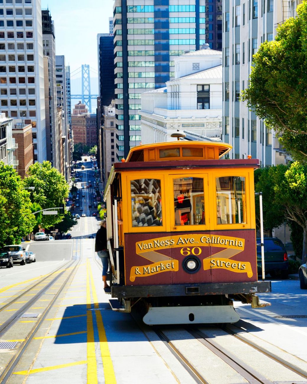 A classic San Francisco cable car travels uphill on a city street with tall buildings and the Bay Bridge visible in the background.