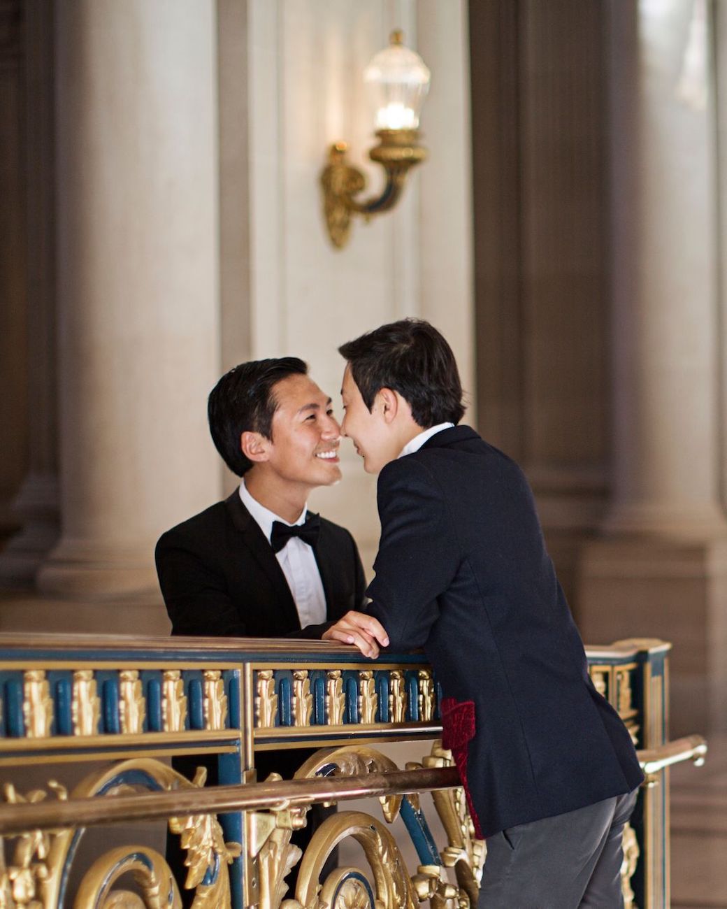 Two men in tuxedos stand close together, facing each other and smiling, on an ornate balcony in an elegant, columned interior.