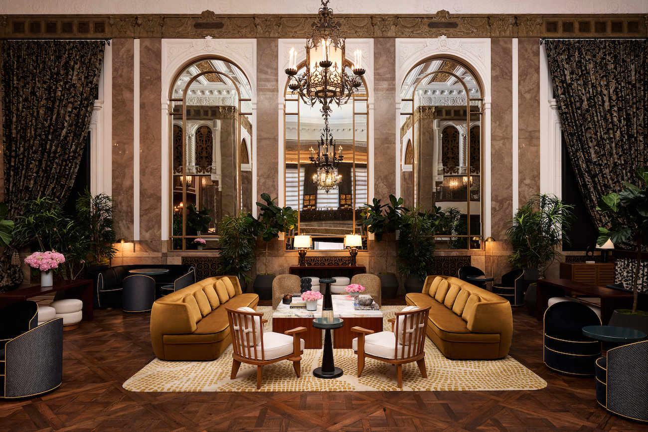 Luxurious hotel lobby with ornate chandeliers, large mirrors, plush brown sofas, white chairs, plants, and a parquet wood floor.