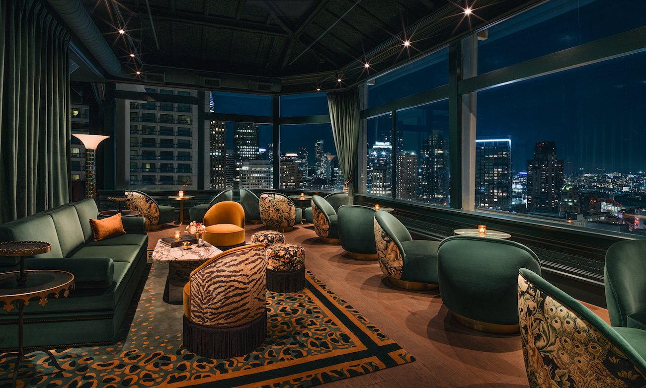 Elegant rooftop lounge with green velvet seating, animal-print accents, and large windows overlooking a city skyline at night.