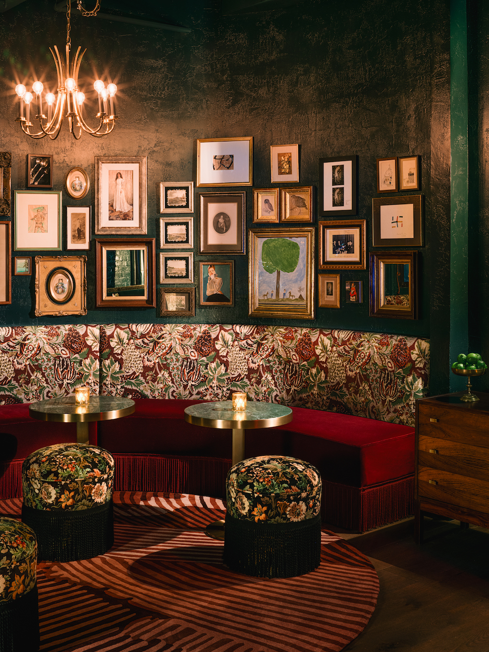 A dimly lit lounge with floral upholstered seating, small round tables, patterned stools, a gallery wall of framed art, and a chandelier overhead.