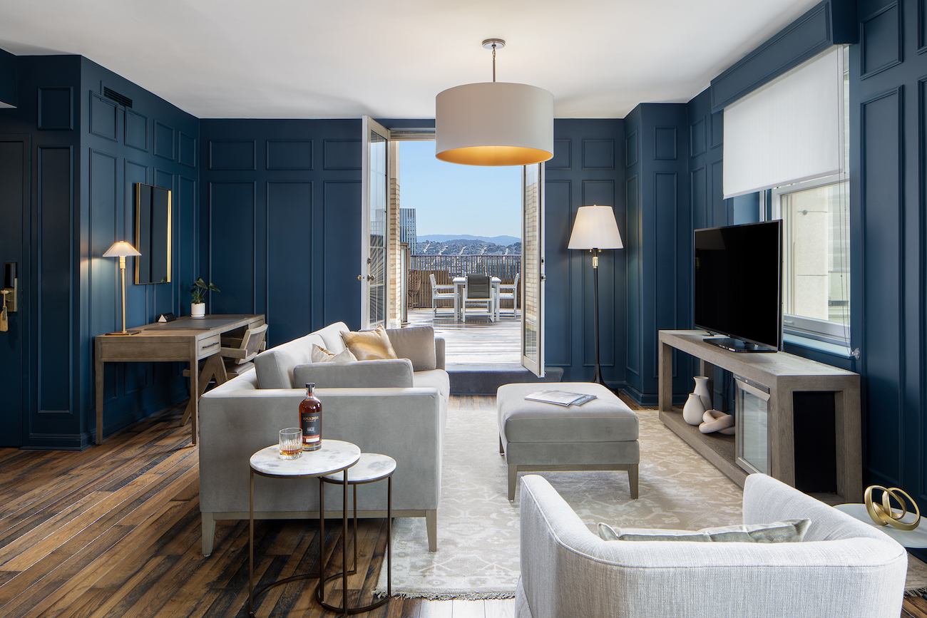 A modern living room with blue paneled walls, gray furniture, a TV, two side tables, desk, and an open door leading to a terrace with outdoor seating.