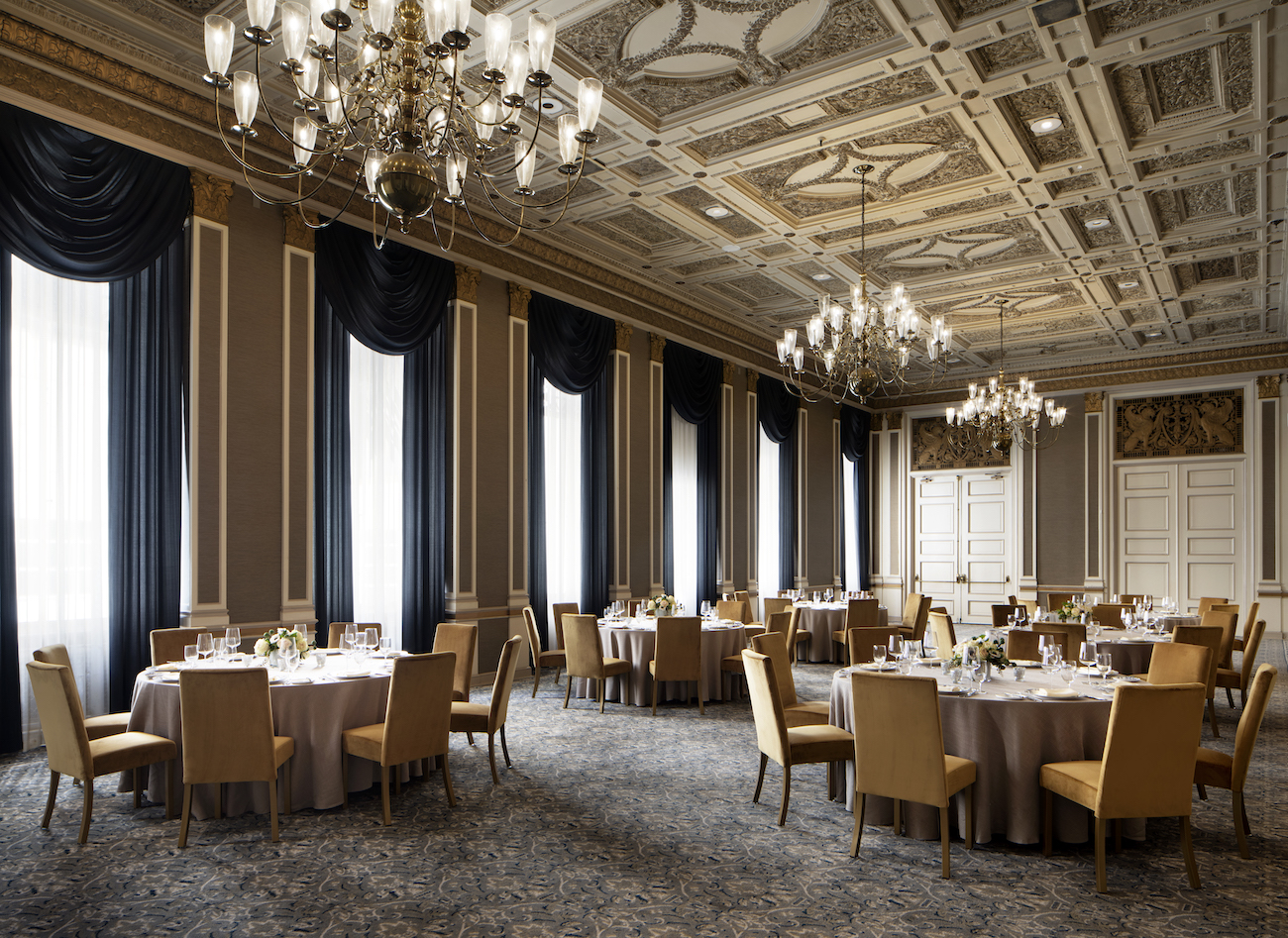 Elegant banquet hall with round tables set for a formal event, featuring chandeliers, ornate ceiling, large windows with dark drapes, and beige walls.