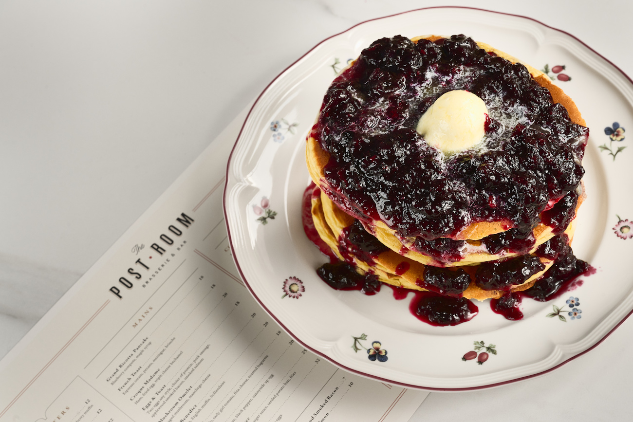 A stack of pancakes topped with blueberry compote and a pat of butter on a floral plate, placed next to a printed restaurant menu.