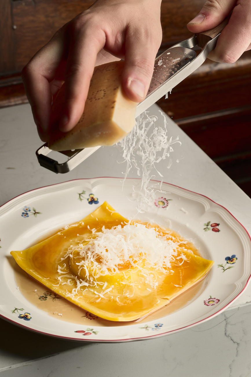 A hand grates cheese over a large raviolo with a runny yolk center on a decorative plate.