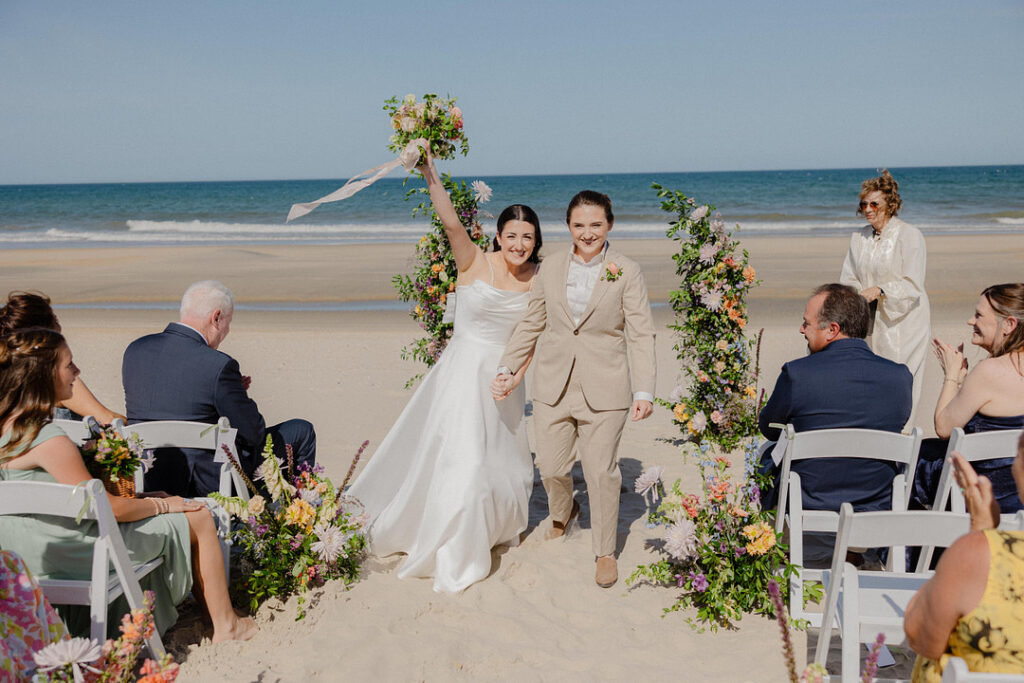 Two women, one in a white dress and one in a tan suit, walk hand-in-hand down a beach aisle lined with flowers, celebrating after their wedding ceremony.
