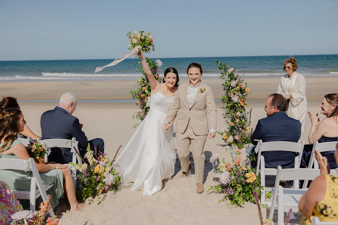 Two women, one in a white dress and one in a tan suit, walk hand-in-hand down a beach aisle lined with flowers, celebrating after their wedding ceremony.