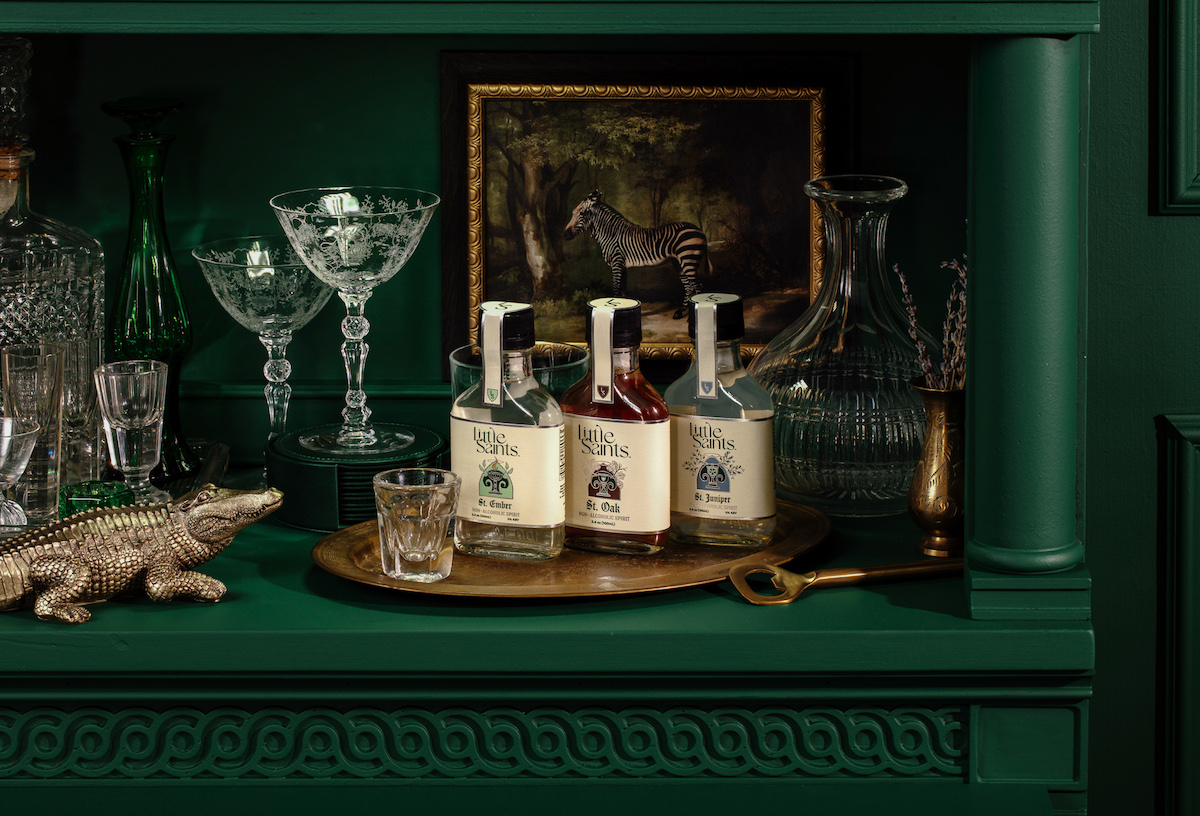 Three bottles of spirits on a gold tray sit among glassware, a crocodile figurine, and a framed animal painting on a green shelf.