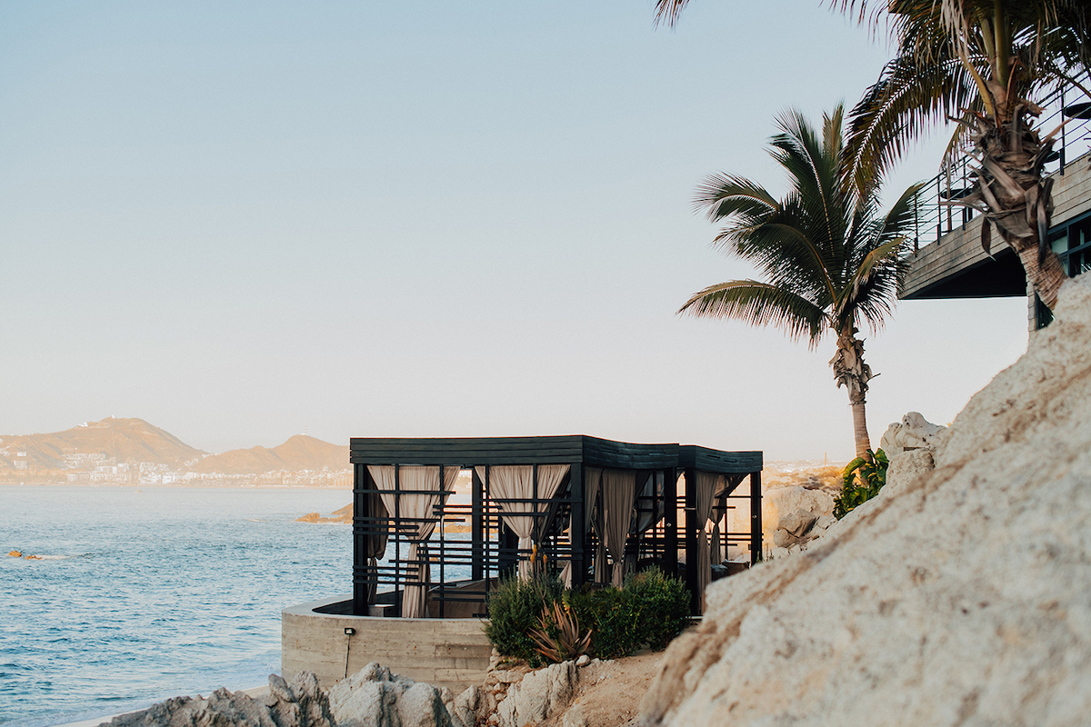 Modern gazebo structure with glass walls overlooking the ocean, situated on a rocky shoreline with palm trees and distant hills in the background.