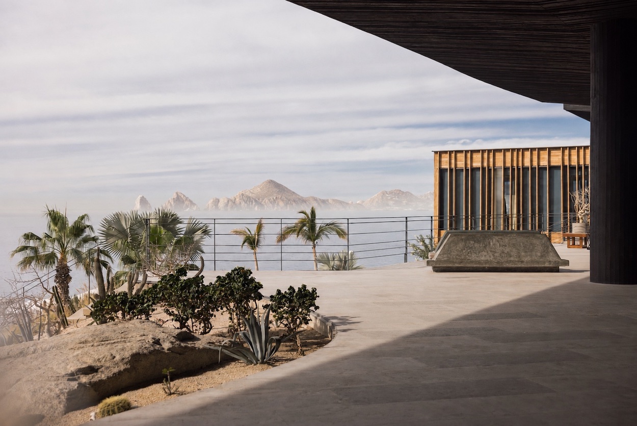 Modern patio with desert plants, a wooden structure, and a view of the ocean and distant mountains under a partly cloudy sky.