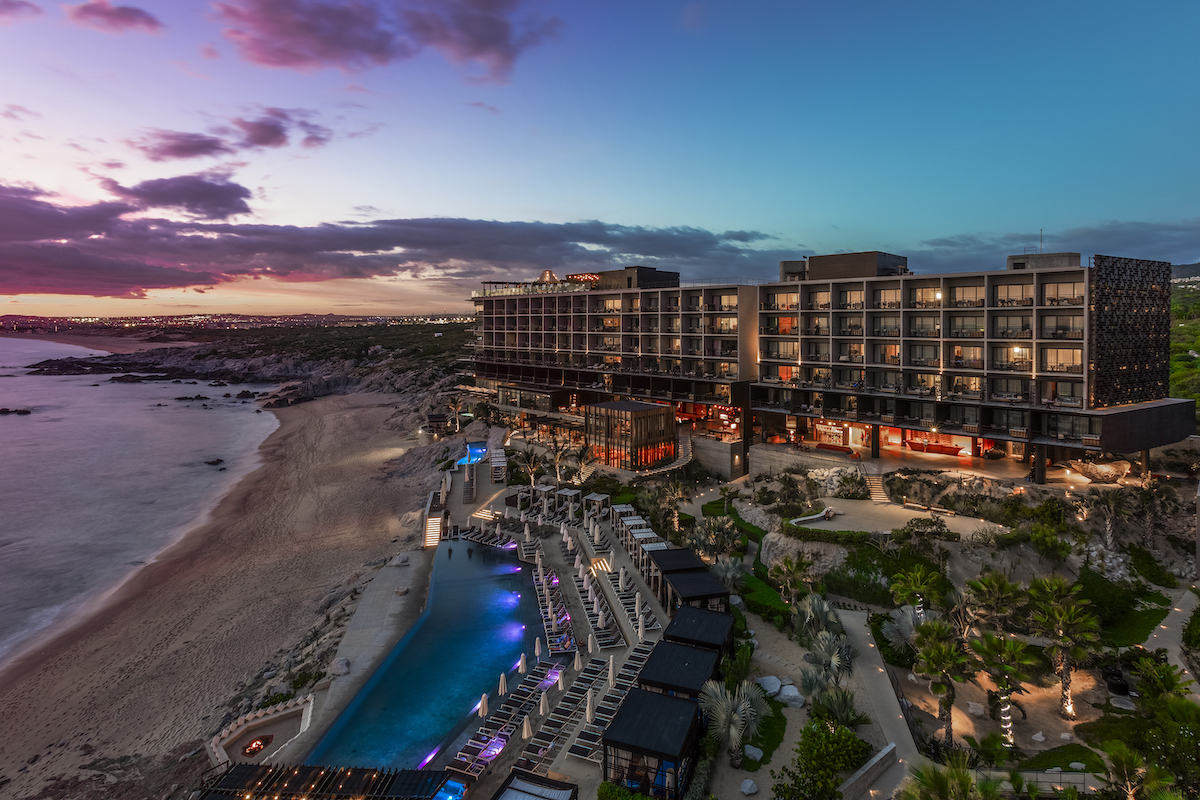 A modern beachfront resort with illuminated pool and lounge areas sits beside the ocean at sunset, with colorful sky and palm trees visible.