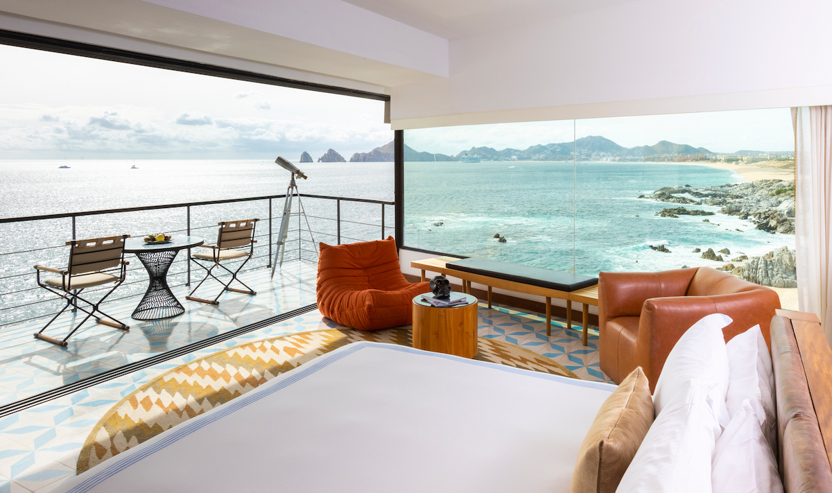 Modern hotel room with large windows and a balcony overlooking the ocean, featuring a telescope, seating area, and scenic coastal view.