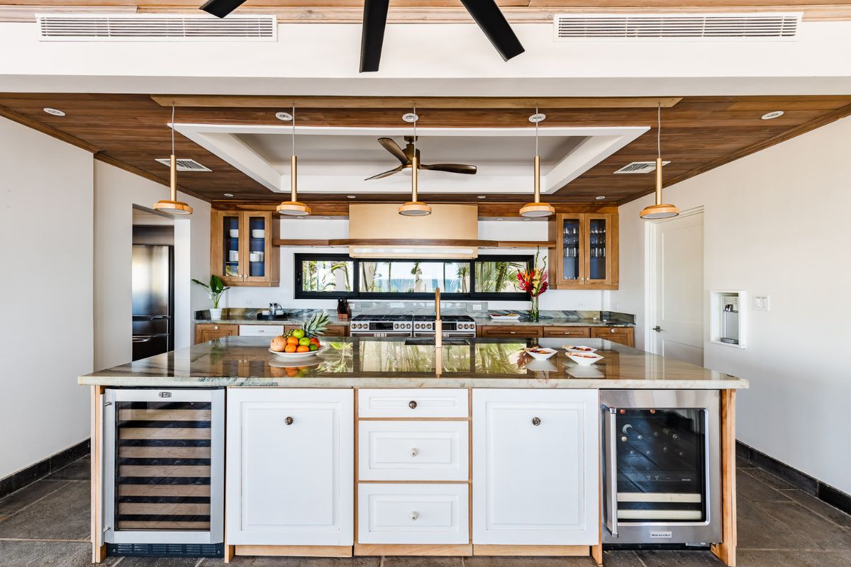 Modern kitchen with a large island featuring a wine fridge, pendant lights, stainless steel appliances, and wooden accents on the ceiling and cabinets.