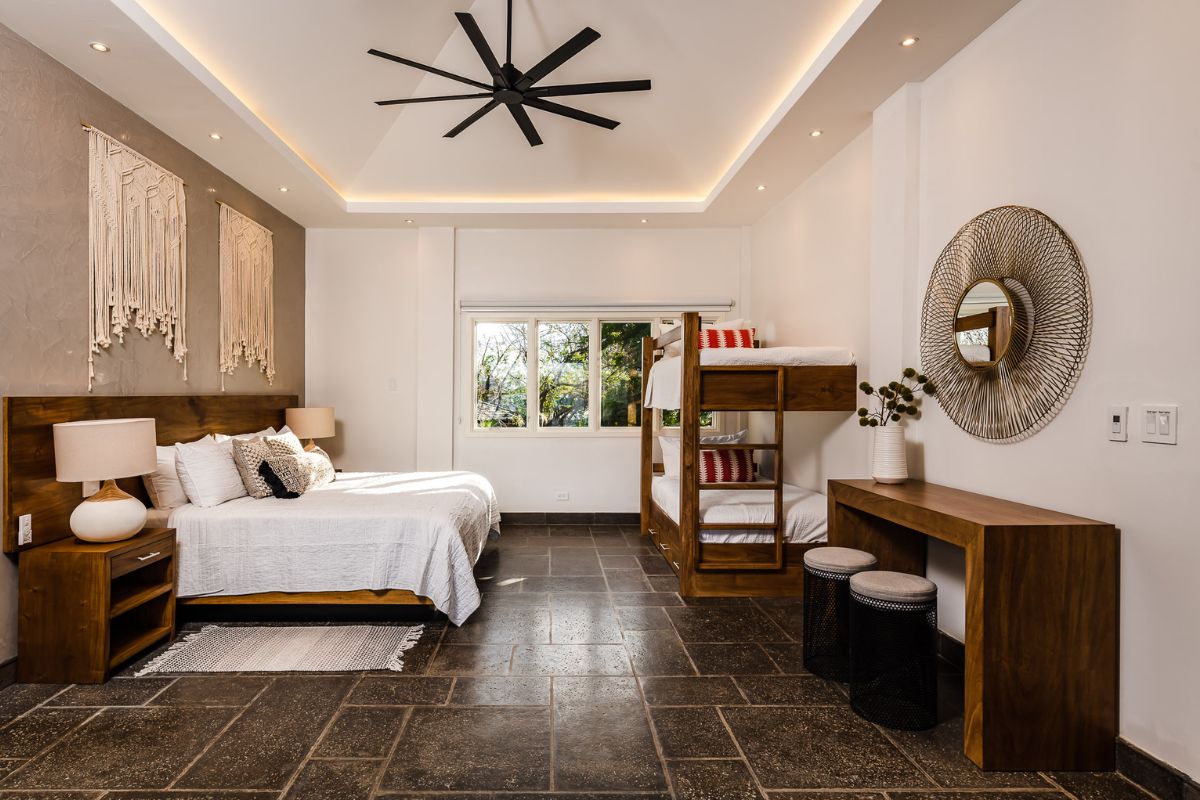 Modern bedroom with a double bed, bunk beds, wooden furniture, a large ceiling fan, and a window overlooking greenery.