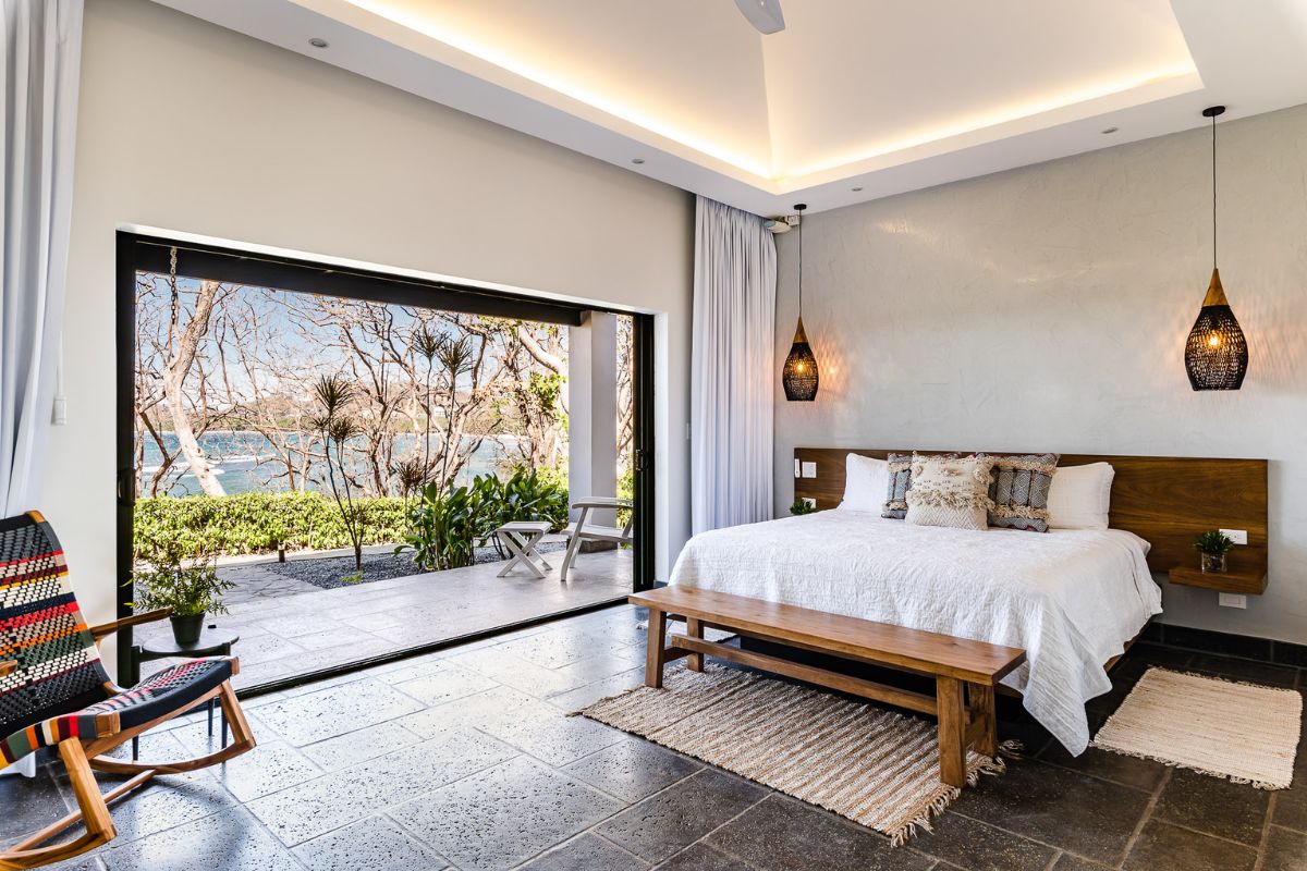 Modern bedroom with large sliding glass door open to a patio and garden view, featuring a wooden bed, bench, decorative chair, and pendant lights.
