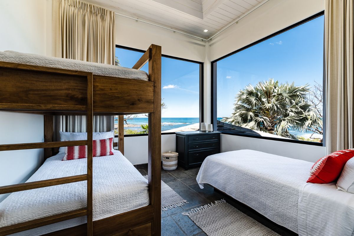 Bedroom with a wooden bunk bed and a single bed, large windows showing a view of the ocean and palm trees, and a dresser by the windows.