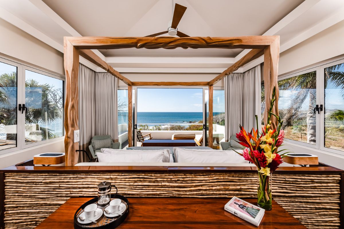 A modern bedroom with a wooden canopy bed, large windows showing an ocean view, a vase of tropical flowers, and a tray with tea set on a wooden table.