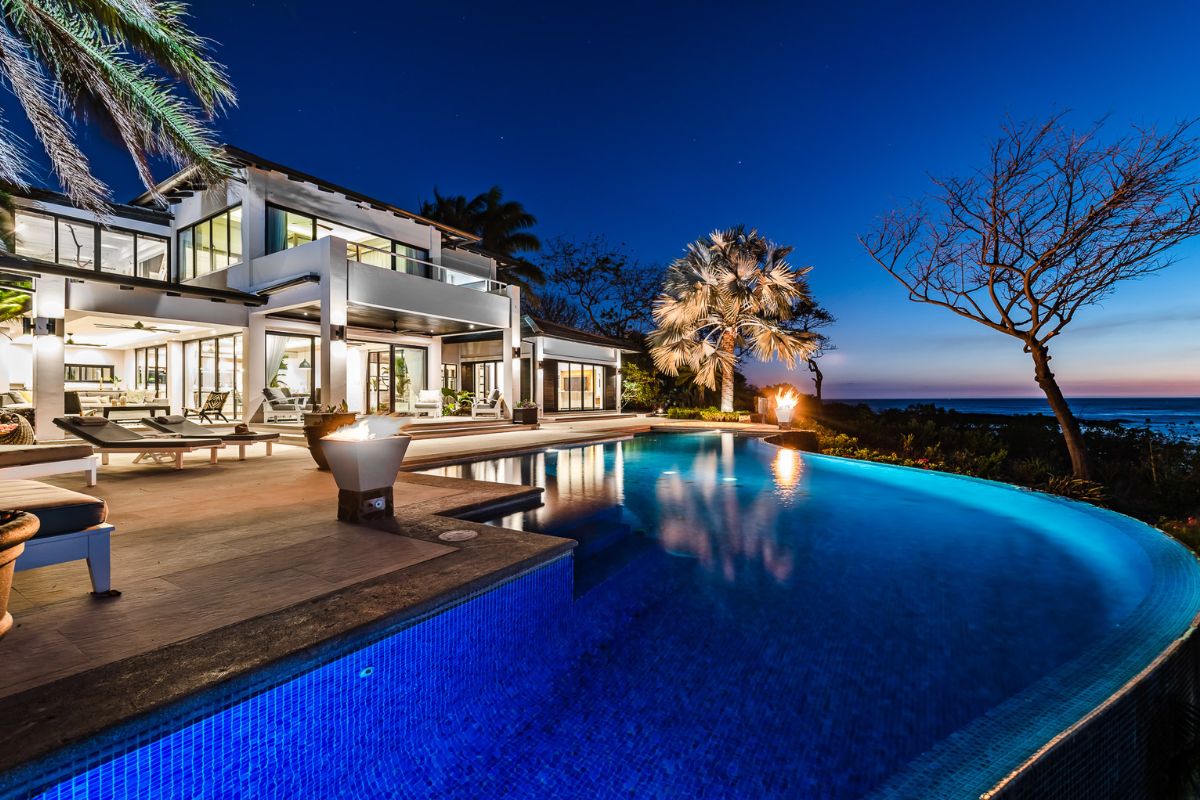 Modern luxury house with large windows and an illuminated infinity pool overlooking the ocean at dusk, surrounded by palm trees and outdoor seating areas.