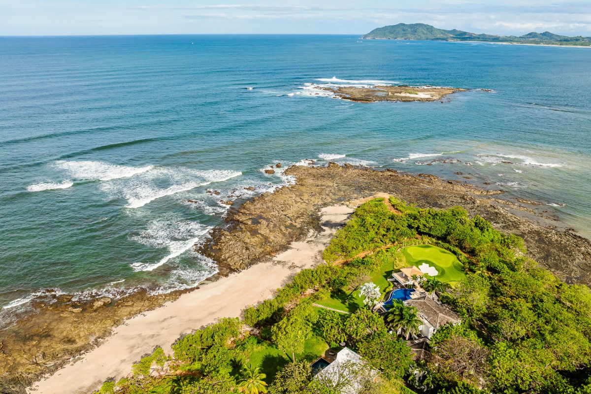 Aerial view of a rocky coastline with waves, sandy beach, lush green vegetation, and a distant island under a blue sky.