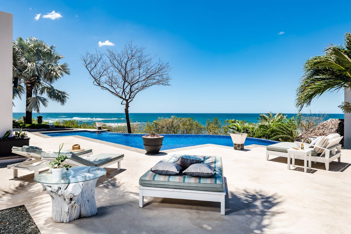 Outdoor patio with lounge chairs, a small pool, and palm trees overlooking the ocean under a clear blue sky.