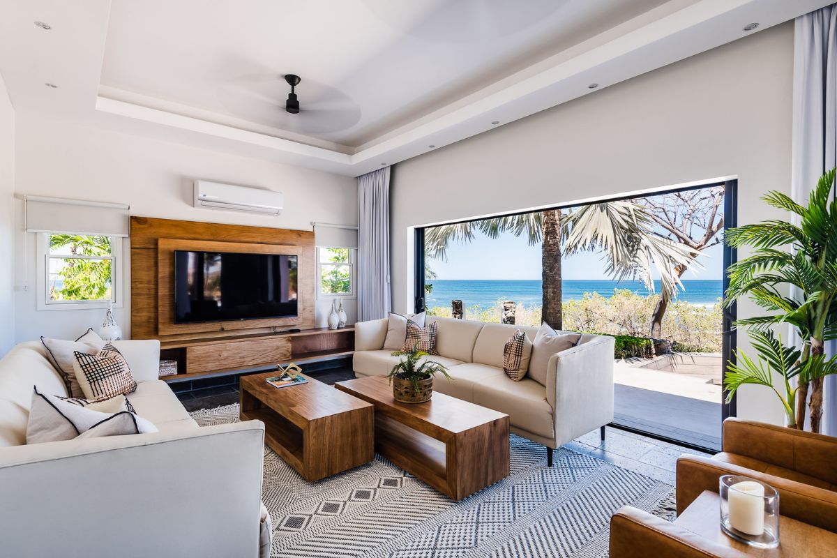 Modern living room with white sofas, wooden furniture, and large windows opening to a view of palm trees and the ocean.