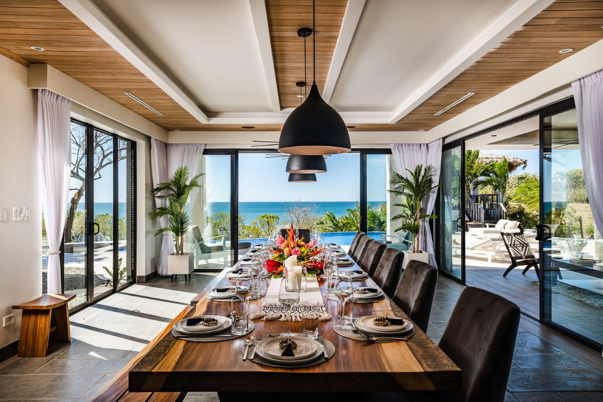 A modern dining room with a long wooden table set for a meal, large windows, ocean view, and contemporary decor.