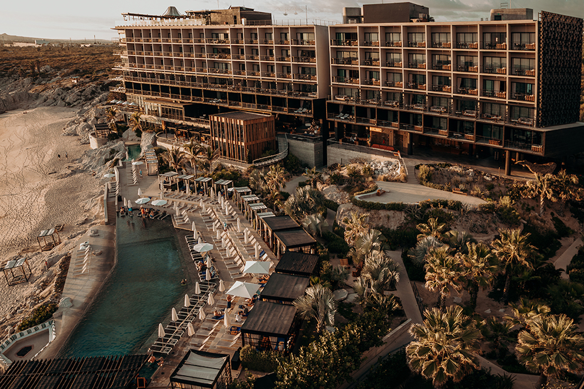 Aerial view of a large beachfront hotel with multiple floors, a long outdoor pool, lounge chairs, palm trees, and guests relaxing by the water.