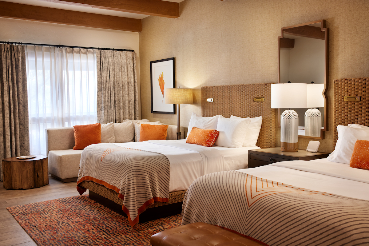 Hotel room with two double beds, a sofa, side tables, lamps, a large window with curtains, and neutral decor accented with orange pillows and blankets.