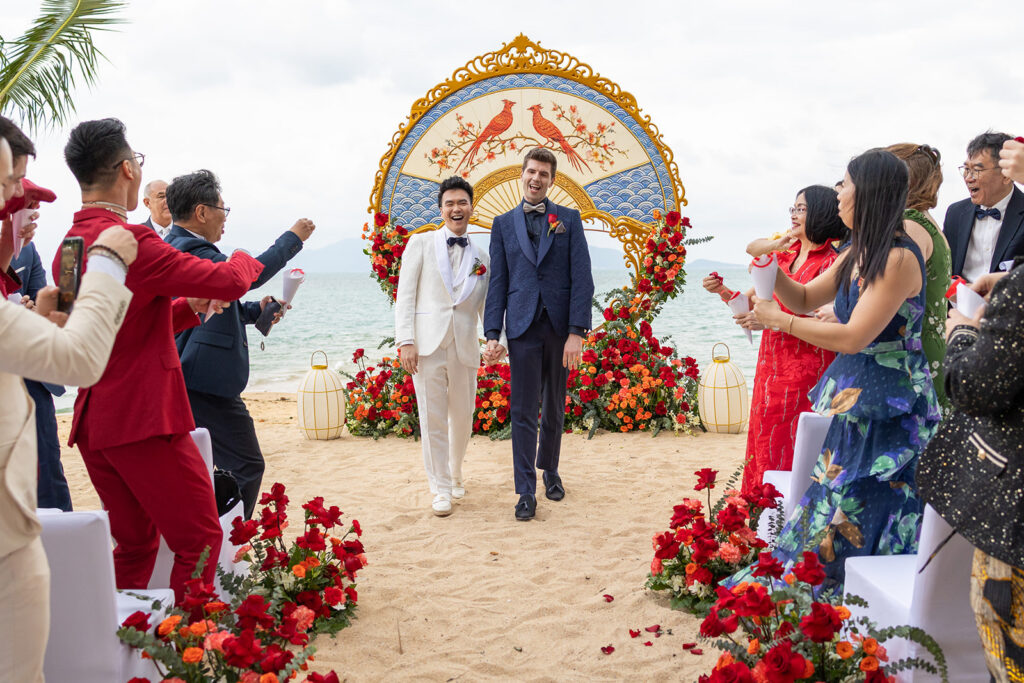 Two grooms walk hand in hand down an aisle on a beach, surrounded by guests and red floral decorations, with a decorative arch in the background.