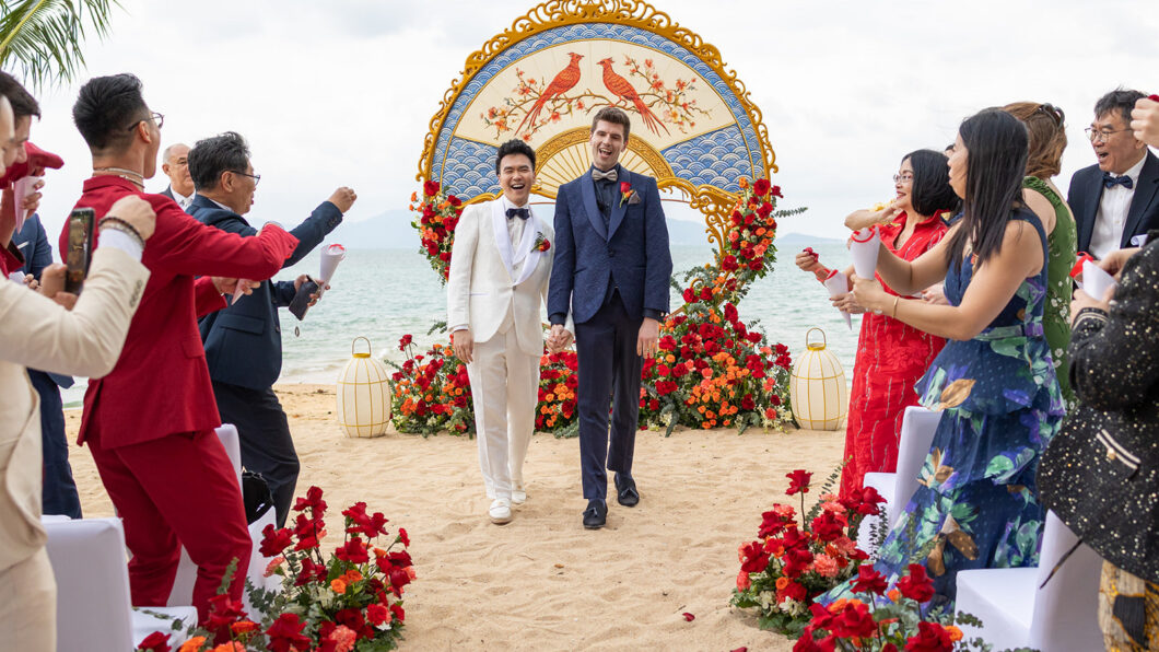Two grooms walk hand in hand down an aisle on a beach, surrounded by guests and red floral decorations, with a decorative arch in the background.