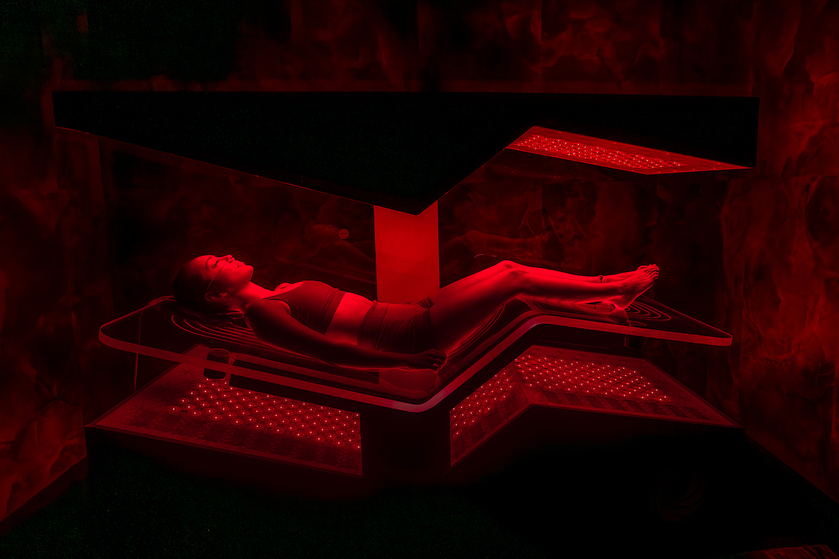 A person in swimwear lies on a platform under red lighting in a dark room, possibly undergoing a light or heat therapy treatment.