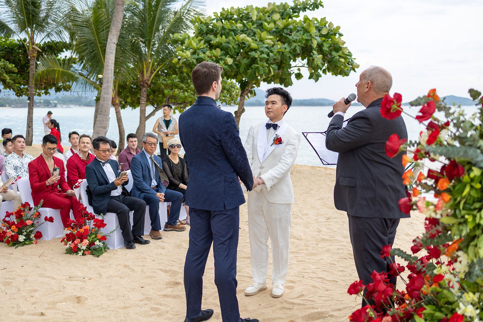 Two grooms stand facing each other on a beach as an officiant speaks; guests are seated nearby, some in red suits, with floral decorations around.