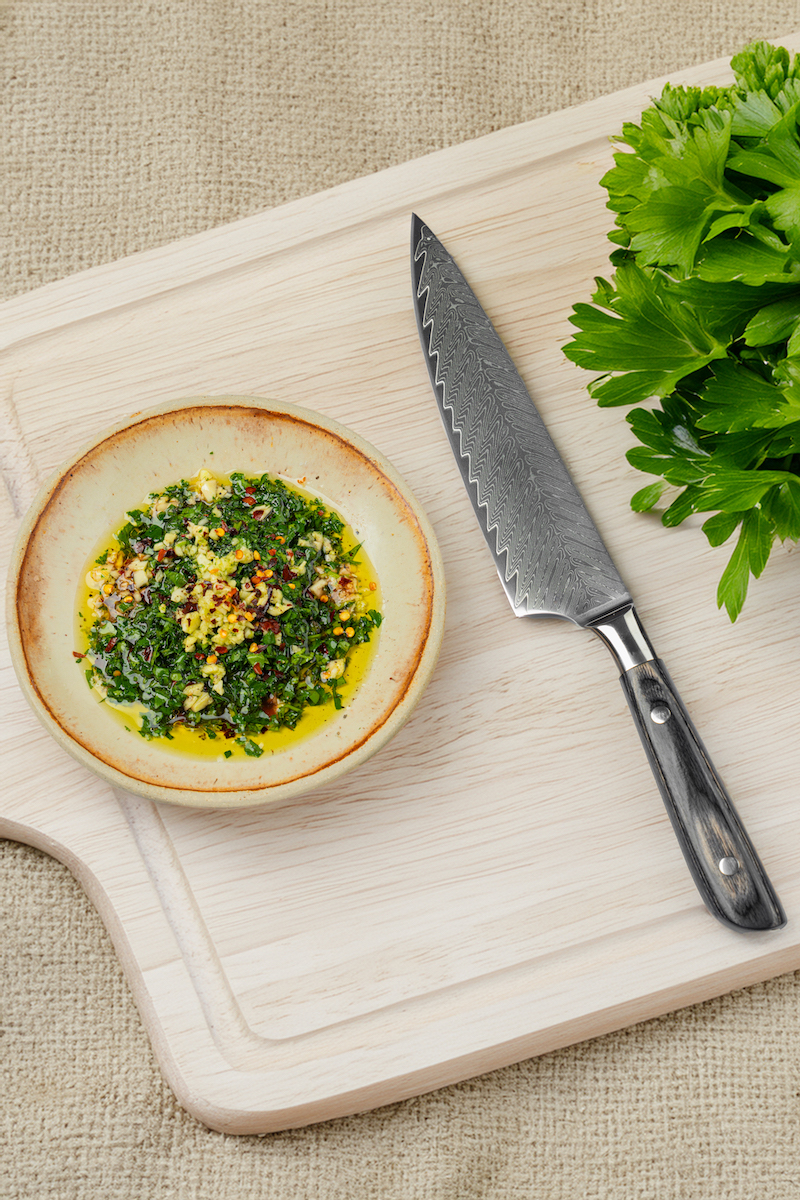 A bowl of chopped herbs and spices in oil sits on a wooden cutting board next to a chef’s knife and a bunch of fresh parsley.