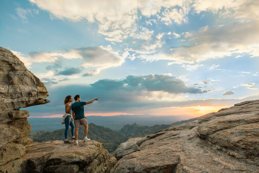 Two people stand on a rocky cliff at sunset, with one person pointing toward the horizon; mountains and a dramatic sky are visible in the background.