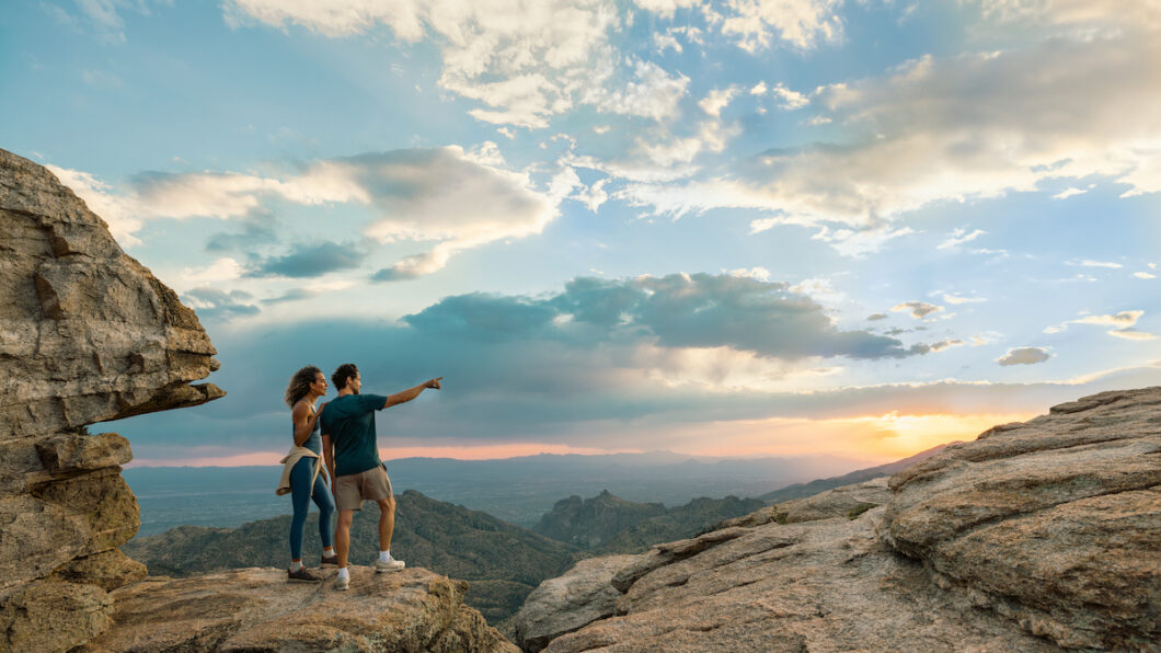 Two people stand on a rocky cliff at sunset, with one person pointing toward the horizon; mountains and a dramatic sky are visible in the background.