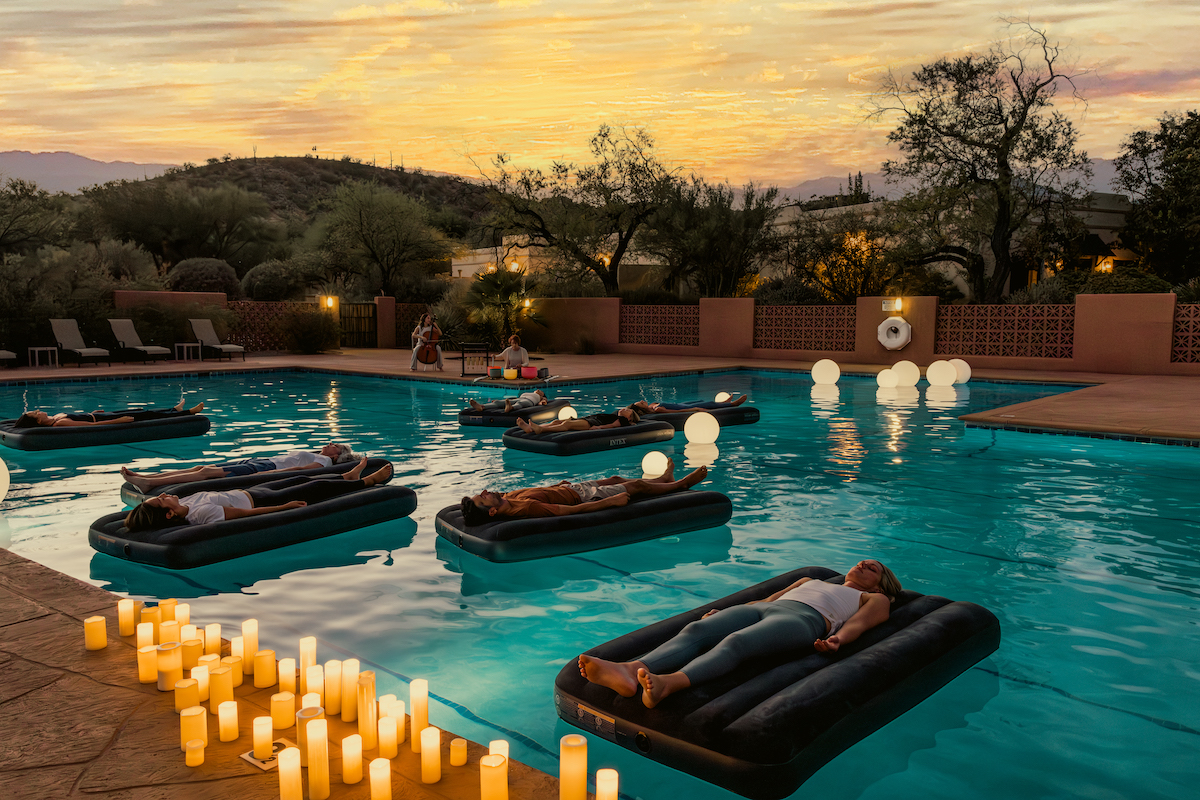 People are lying on floating air mattresses in a pool at sunset, surrounded by lit candles and glowing orbs, with trees and mountains in the background.