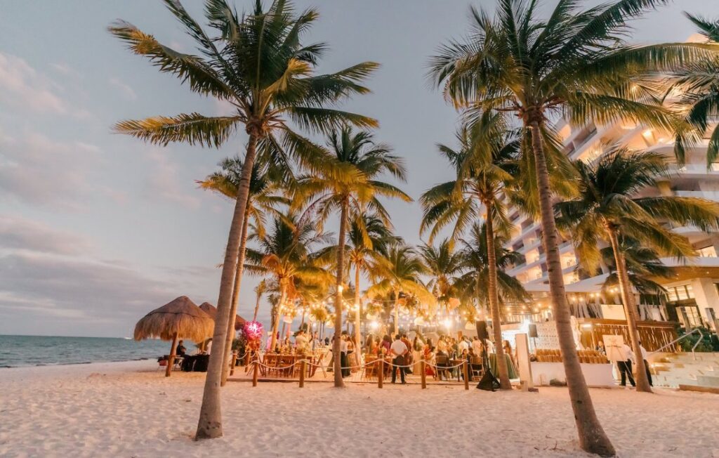 Outdoor beach event at sunset with people gathered under palm trees and string lights, near a thatched hut and a multi-story building.