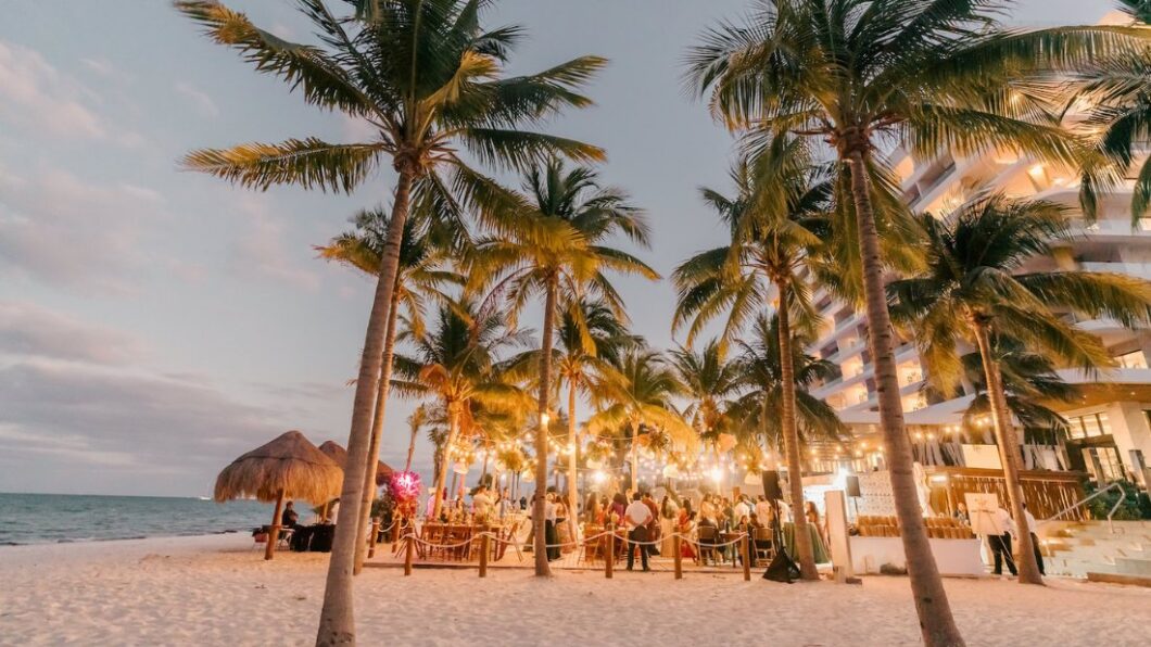 Outdoor beach event at sunset with people gathered under palm trees and string lights, near a thatched hut and a multi-story building.