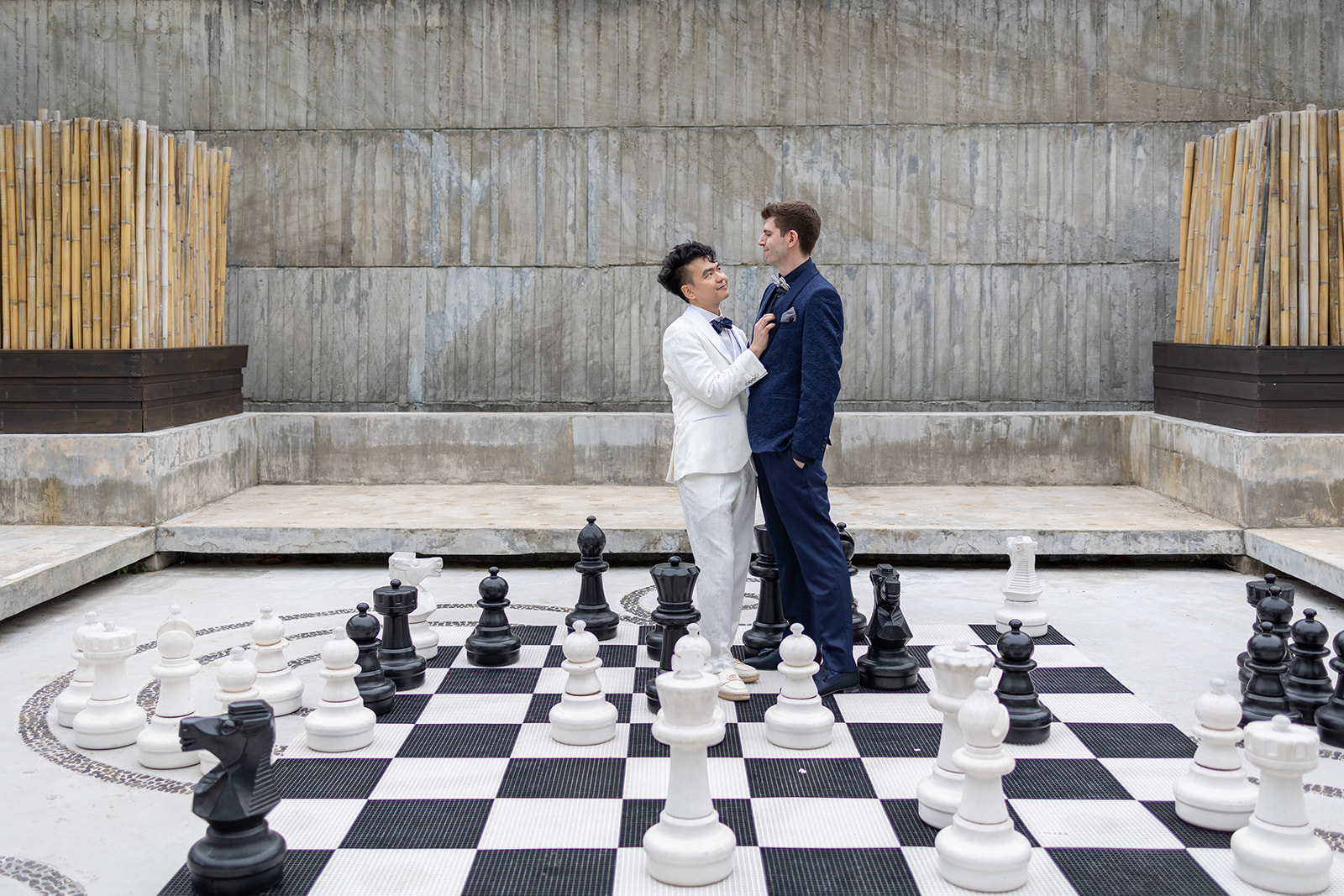 Two men in suits stand face to face on an outdoor life-size chessboard, surrounded by large chess pieces and concrete walls.