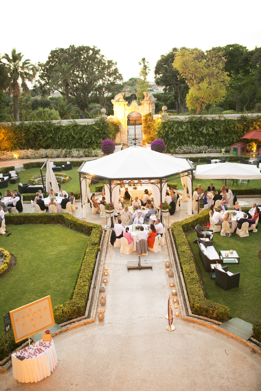 Outdoor event with guests seated at round tables under a white canopy, surrounded by gardens and greenery at sunset.