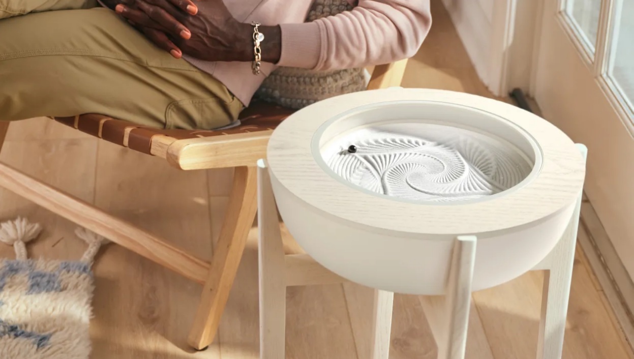 A person sits in a chair next to a white, circular tabletop sand art device with geometric patterns drawn in the sand.